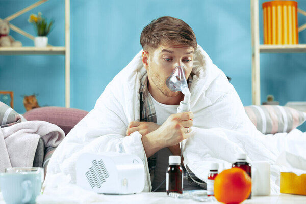 Bearded sick man with flue sitting on sofa at home covered with warm blanket and using an inhaler when coughing. The illness, influenza, pain concept. Relaxation at Home. Healthcare Concepts.