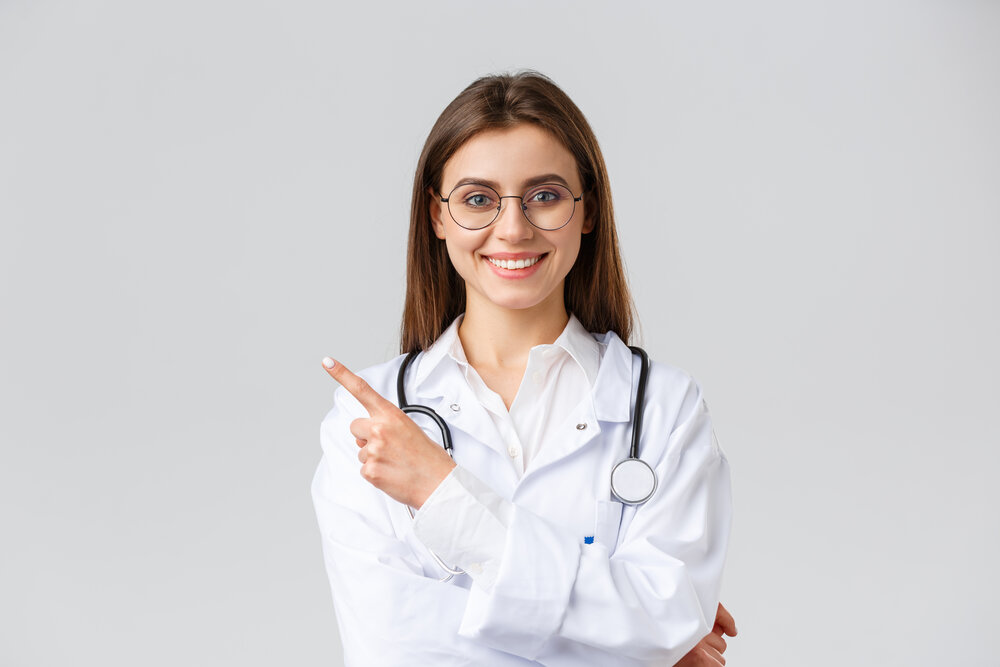 Dobre vyzerajúca profesionálna lekárka v bielom plášti a okuliaroch, ukazuje prstom doľava, priateľsky sa usmieva, ukazuje informácie..