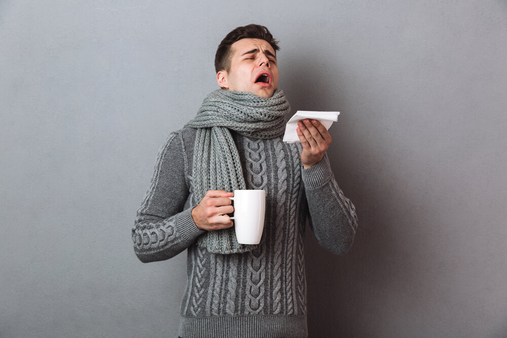 Sick Man in sweater and scarf sneezes while holding cup of tea over gray background