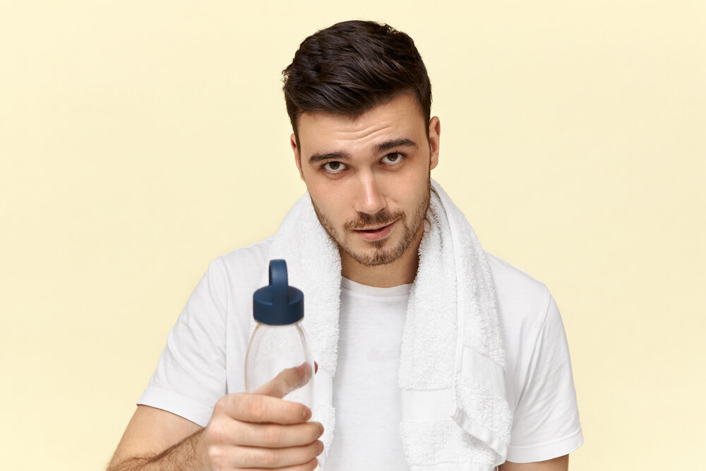 pekný mladý európsky muž s bielym uterákom okolo krku odporúča pitie vody z plastového pohára 