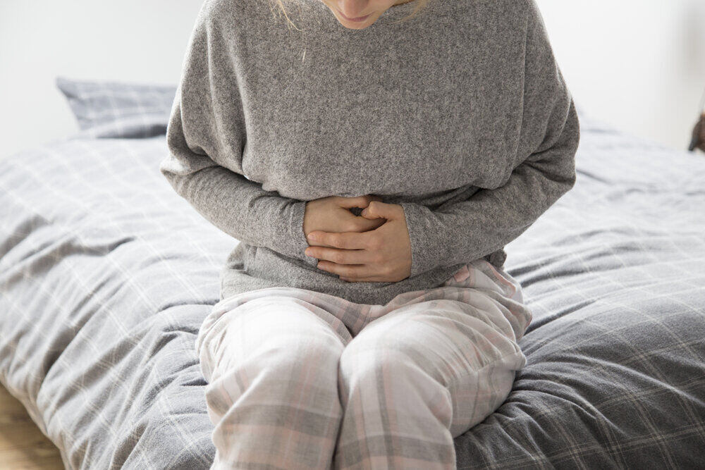 Chorá žena v sivom domácom oblečení sedí na posteli a drží si ruky na bruchu.