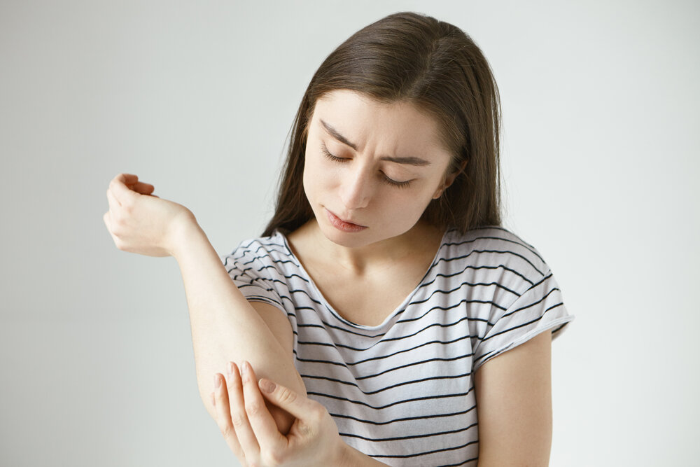 obrázok frustrovanej mladej tmavovlasej ženy študujúcej pokožku na ruke po tom, čo na nej objavila kliešťa
