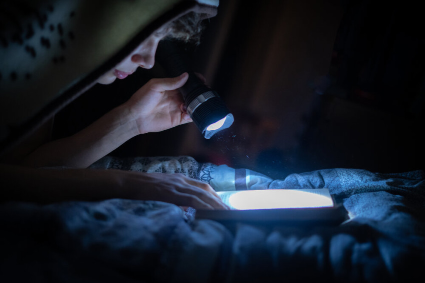 mladý človek s baterkou si číta knihy v noci v posteli
