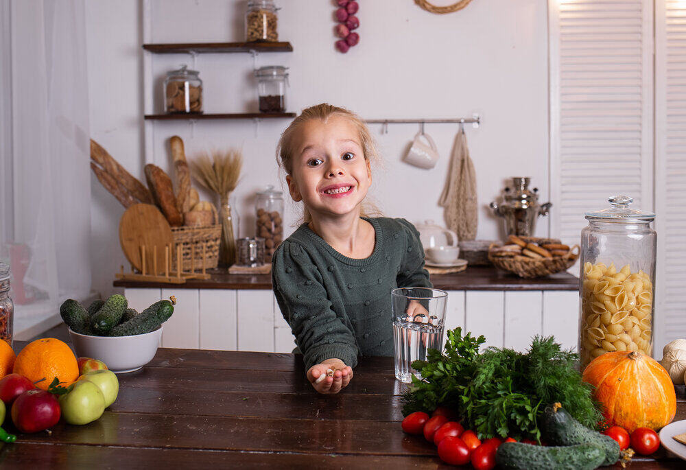 šťastná malá slečna sedí pri stole s pohárom vody a vitamínmi v ruke
