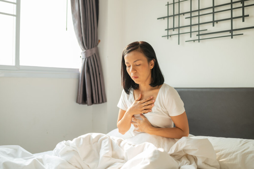 Ranná nevoľnosť. Mladá tehotná žena sediaca na posteli, zakrývajúca si ústa a pocit nevoľnosti počas tehotenstva, Žena v bielom pyžame trpiaca kyslým refluxom, keď sa ráno prebúdza na posteli.