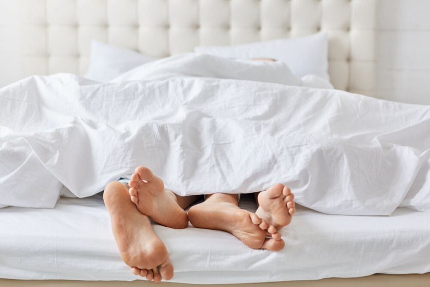 záber nôh páru pod bielou posteľnou bielizňou v spálni, ako si doma užívajú